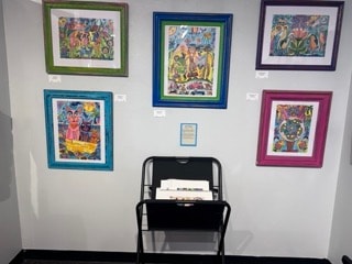 Five framed folk art paintings displayed on a gallery wall with a folding chair