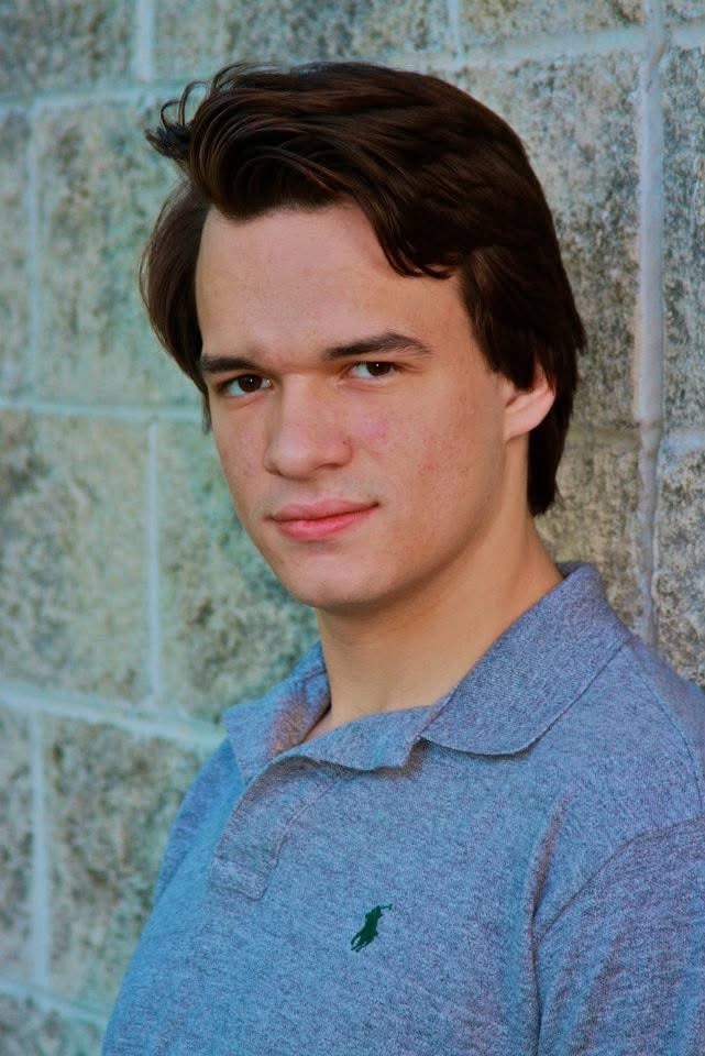Young man in grey polo shirt leaning against a stone brick wall